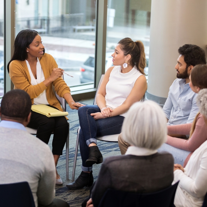 A group of diverse people in a circle listening to a mentor leading a verbal exercise while taking notes 