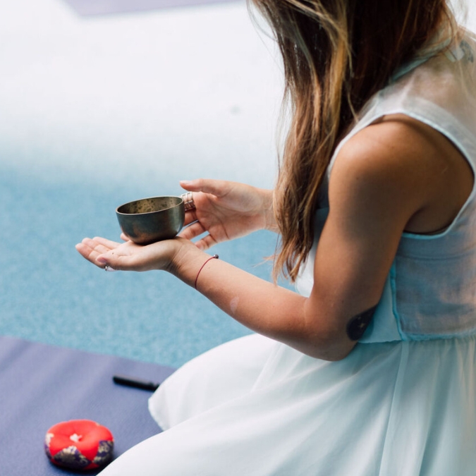 A young woman pictured in silhouette holding a meditation bell outside while sitting comfortably on her knees on a yoga mat