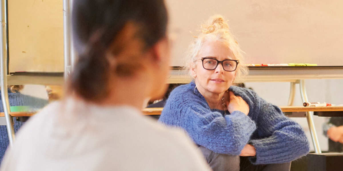 Blonde-haired facilitator wearing glasses and a blue sweater listening to youth during a session